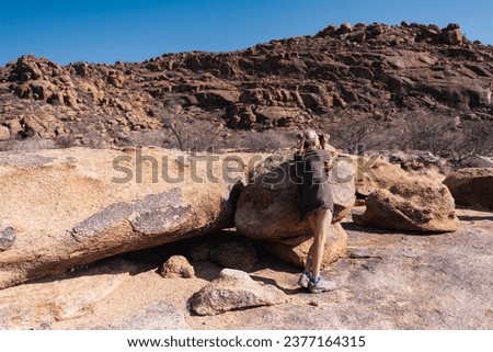 A woman looks through a pair of binoculars while leaning on a large rock in the Erongo Region on Namibia