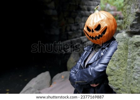 An adult model wearing a pumpkin head mask poses for a portrait by leaning against the stones of the Glen Span Arch with crossed arms