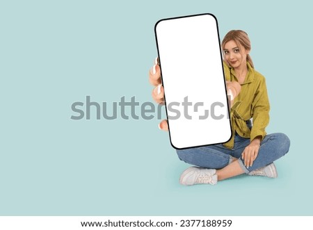Phone with blank screen, full length body view sitting woman hold mobile phone with blank screen. Showing big smartphone mock up to camera. Banner advertising design template idea. Isolated blue. Royalty-Free Stock Photo #2377188959