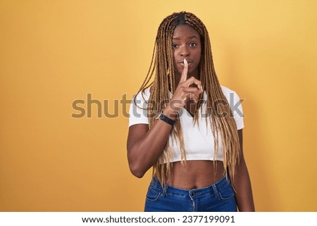 African american woman with braided hair standing over yellow background asking to be quiet with finger on lips. silence and secret concept. 