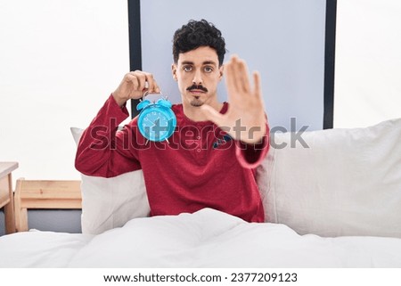 Hispanic man holding alarm clock in the bed with open hand doing stop sign with serious and confident expression, defense gesture 