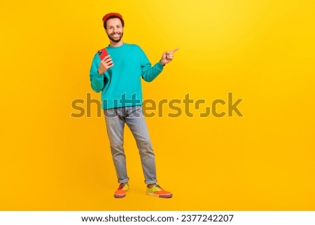 Full size photo of handsome guy wear teal sweatshirt hold smartphone indicating at offer empty space isolated on yellow color background