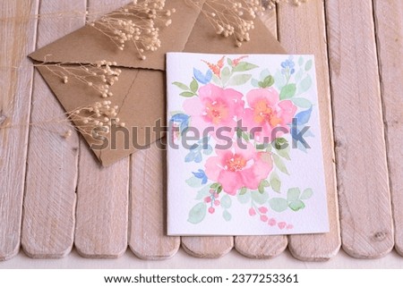 Wedding invitation hand painted watercolor floral card romantic style pink flowers green blue colors leaves brown craft envelope on wood background