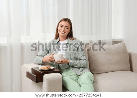 Happy woman holding cup of drink on sofa armrest wooden table at home Royalty-Free Stock Photo #2377254141