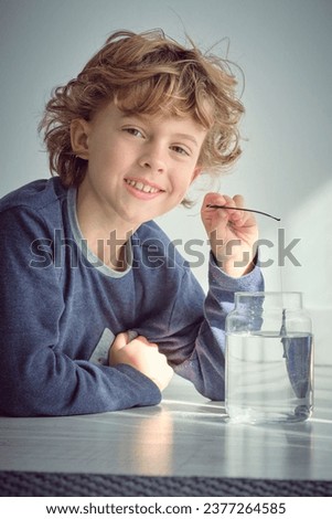 Positive little kid with curly blond hair putting toy small fishing rod into transparent glass jar of water while looking at camera against white background