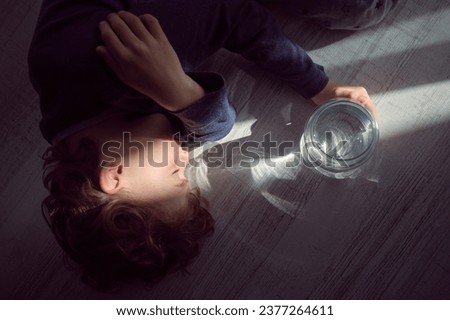 Overhead of little kid with curly hair lying peacefully on floor in sun ray shining through glass bottle with water