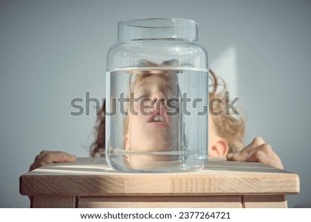 Napping kid with blond hair and open mouth near glass jar of water placed on wooden stool against white background