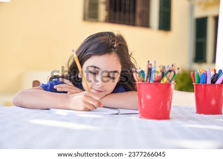 Adorable creative black haired girl drawing on paper of copybook with pencils at table at daytime alone