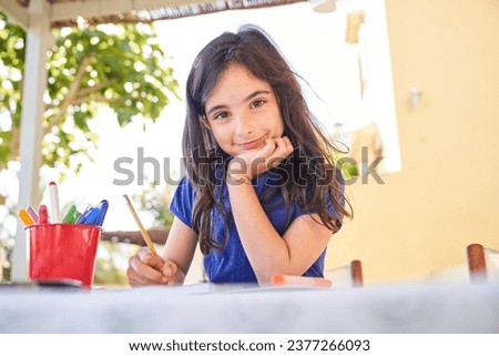 Smiling little hispanic girl in casual clothes looking at camera while sitting at table and drawing pictures with colorful pens