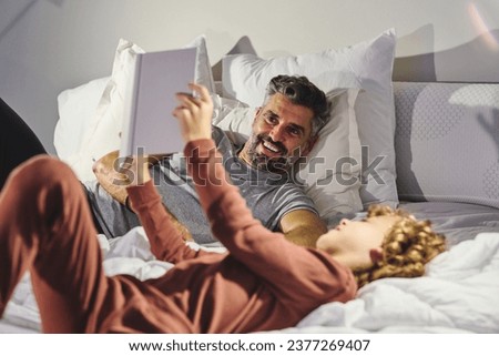 Cheerful man and boy in sleepwear having fun while lying on soft bed and reading interesting book together in cozy bedroom at home Royalty-Free Stock Photo #2377269407