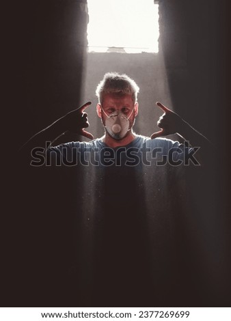 Adult blond haired male surfboard maker in respirator mask showing hawaiian shaka gesture while standing near window illuminated with sunbeam in dark room and looking at camera