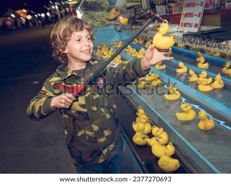 Cute boy in casual clothes catching yellow rubber duck with toy fishing rod while smiling and standing in funfair at night