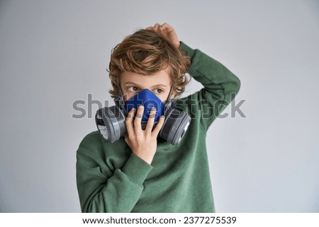 Thoughtful little boy with blond hair in casual clothes putting on protective respirator and looking away against gray background Royalty-Free Stock Photo #2377275539
