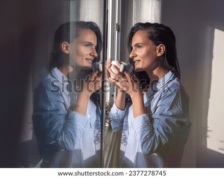 Side view of charming female with long dark hair and in pajamas drinking hot coffee from cup while standing near window in sunny morning Royalty-Free Stock Photo #2377278745
