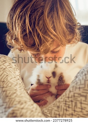 Adorable tender boy in pajama caressing fluffy rabbit with white fur and spots while sitting in light room at home