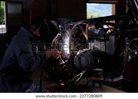 Side view of faceless mechanic wearing protective helmet and welding motorcycle in dark workshop Royalty-Free Stock Photo #2377280809