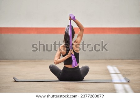 Back view of unrecognizable female athlete in activewear sitting on gray sports mat and doing exercises with elastic band during training in gym Royalty-Free Stock Photo #2377280879