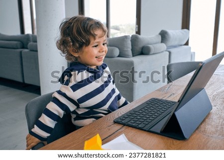 Side view of little boy in casual clothes sitting at table with tablet and having fun while watching funny cartoon at home