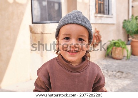Cute funny little boy in casual clothes and hat smiling and looking at camera standing in yard near aged house on sunny day