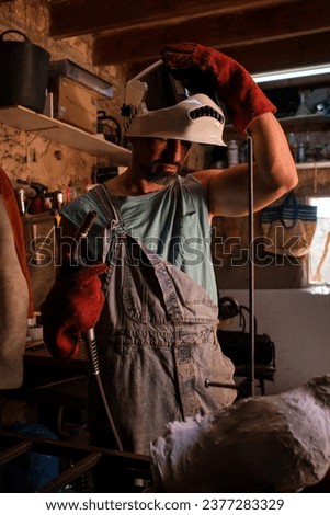 Serious craftsman in uniform with gloves putting on protective helmet and looking down while standing at workbench with welding torch during workday in professional workshop Royalty-Free Stock Photo #2377283329