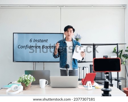 Asian life coach teaching about 6 Instant Confidence Boost and share course on tv screen while selfie record lesson with smartphone and tripod for client watch online later in education studio room Royalty-Free Stock Photo #2377307181