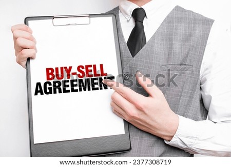 BUY-SELL AGREEMENT inscription on a notebook in the hands of a businessman on a gray background, a man points with a finger to the text