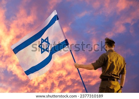 Israeli soldier with Israel flag against a fiery sunset. Remembrance Day - Yom HaZikaron, Patriotic holiday, Israeli Independence Day - Yom Ha'atzmaut concept. Royalty-Free Stock Photo #2377310189