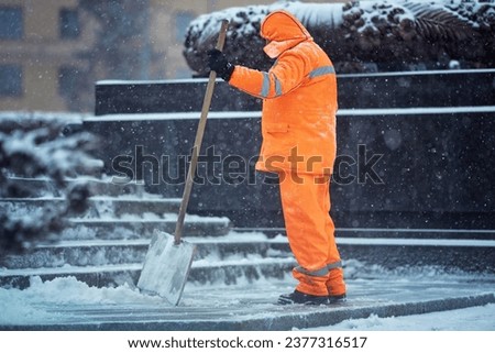 Utility man with snow shovel clear snow during blizard, snow removal work. Municipal worker in uniform with snowshovel in hands clean snowy sidewalk and walkway during winter storm Royalty-Free Stock Photo #2377316517