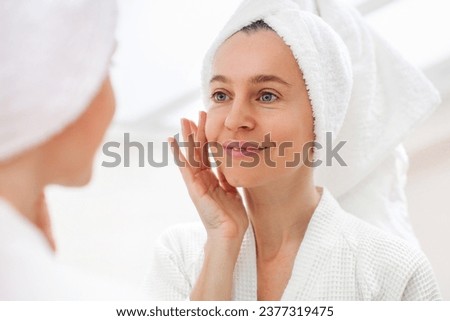 Happy 40s middle aged woman model touching face skin looking in mirror reflection. Smiling adult lady pampering, healthy moisturized skin care, aging beauty, skin care treatment cosmetics concept Royalty-Free Stock Photo #2377319475
