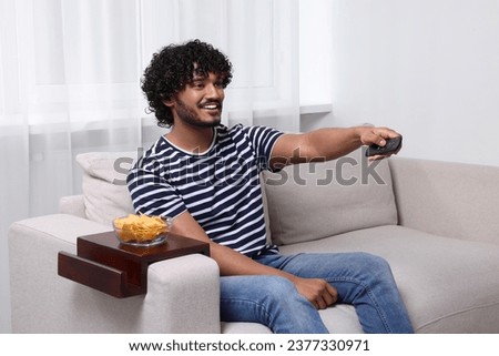 Happy man switching TV channels at home. Nacho chips on sofa armrest wooden table Royalty-Free Stock Photo #2377330971