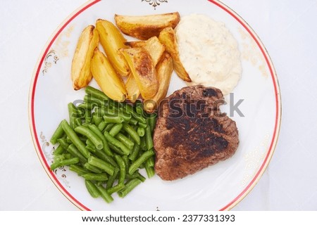 Top view picture on the rustic style porcelain plate with roasted beef steak from tenderloin served with baked potato wedges, green beans and homemade tartar sauce isolated on the white table cloth.