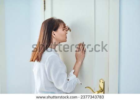 Serious middle-aged woman looking through peephole on front door Royalty-Free Stock Photo #2377343783