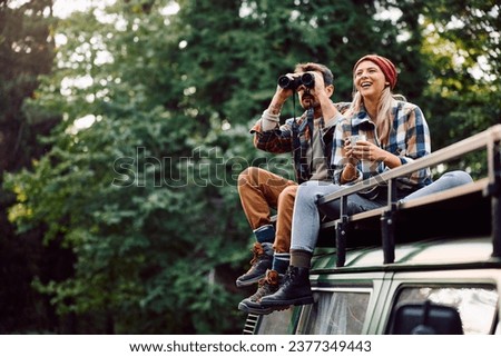 Happy woman and her boyfriend using binoculars while sitting on roof of their camper trailer and spending a day in the woods. Copy space. Royalty-Free Stock Photo #2377349443