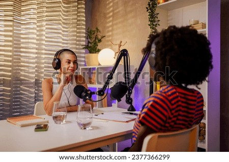 An urban gen z girl is interviewing a guest in her podcast and listening to her while wearing headphones and using cutting-edge technology for recording a podcast. Two girls discussing on the podcast.