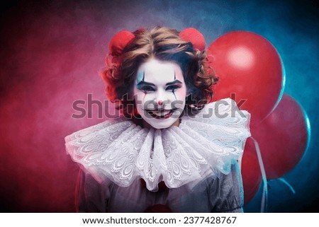 Portrait of a scary clown girl in a white circus costume who smiles widely with red balloons in the background. Scary Halloween.