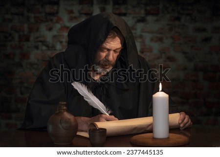 An old monk in his cell writes a message on parchment with a goose quill pen by candlelight. Royalty-Free Stock Photo #2377445135