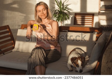 Peaceful middle aged woman enjoys the morning light sitting on the couch at home. Smiling woman is sitting on the sofa with dog and enjoying cup of tea or coffee Royalty-Free Stock Photo #2377486051