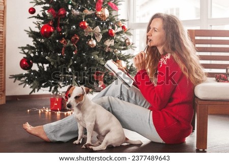 Thinking woman writing list of presents for her family members, sitting at home among Christmas wrapped gift boxes. Dreamy woman taking notes in notebook and smiling