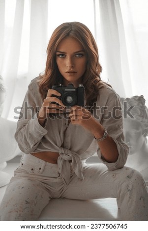 Elegant brunette in light attire with camera, seated among ancient Greek busts on a white table