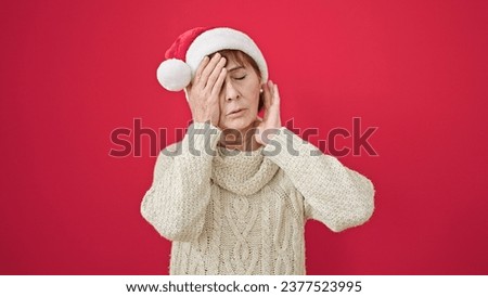 Mature hispanic woman standing with tired expression wearing christmas hat over isolated red background