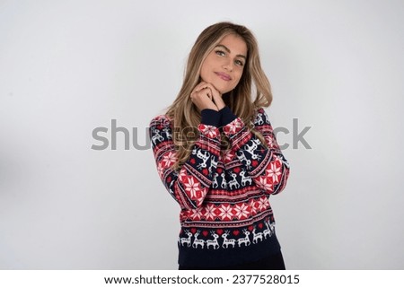 Charming serious Beautiful blonde woman wearing knitted christmas sweater keeps hands near face smiles tenderly at camera
