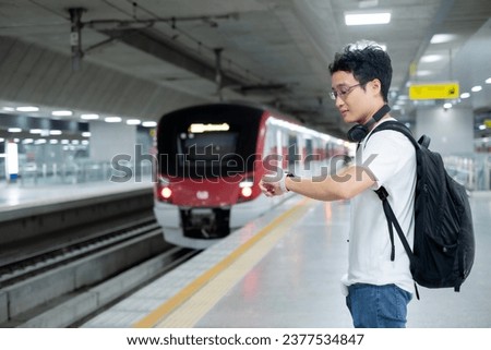 Young Asian millennial man standing in train  while looking at smart watch when the train is coming with black backpack and headphone with happiness.