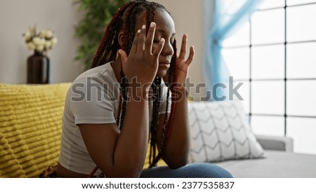 Depressed african american woman with braids sitting alone on sofa at home, unhappy expression reveals headache ache and stress Royalty-Free Stock Photo #2377535837