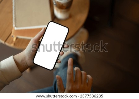 A woman is chilling in a cafe coffee shop and using her smartphone at a table while enjoying her coffee. A white-screen smartphone mockup for display your graphic ads. close-up image Royalty-Free Stock Photo #2377536235