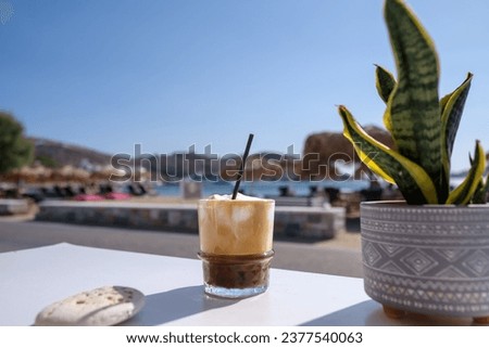 View of an iced Greek  coffee, also known as freddo cappuccino with a straw and the beach of Gialos in Ios Greece

