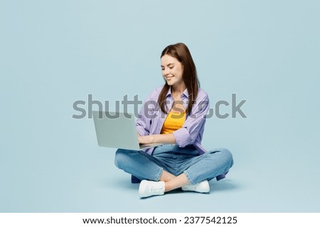Full body young happy IT woman wear purple shirt yellow t-shirt casual clothes sit hold use work on laptop pc computer isolated on plain pastel light blue background studio portrait. Lifestyle concept