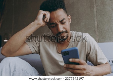 Close up disappointed sad young african american man in beige t-shirt sit on grey sofa indoors apartment using mobile cell phone prop up forehead think rest on weekends stay home Tattoo translate fun. Royalty-Free Stock Photo #2377542197