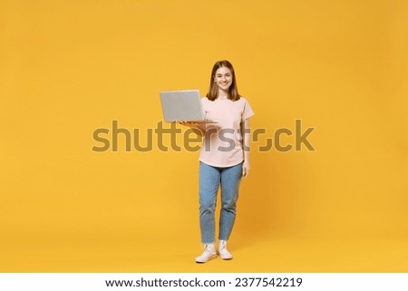 Full length of young caucasian student IT woman 20s in basic pastel pink t-shirt jeans holding laptop pc computer chatting surfing internet looking camera isolated on yellow background studio portrait