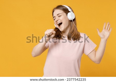 Young fun woman 20s wearing casual basic blank print design pastel pink t-shirt listening to music in headphones sing song in microphone in karaoke isolated on yellow colotbackground studio portrait.