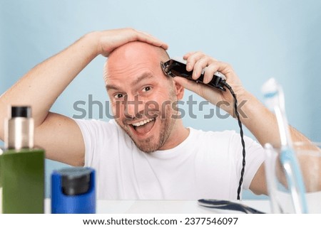Middle aged Caucasian man looks in the bathroom mirror and shaves his head with a hair clipper. Looking extremely happy. Royalty-Free Stock Photo #2377546097